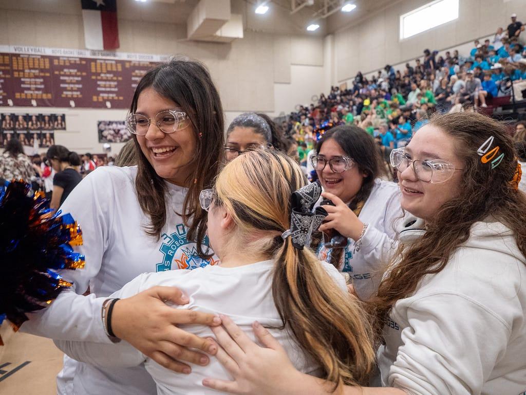 Students in safety glasses hugging and cheering in the stands at the Dripping Springs FIRST Robotics Competition district event